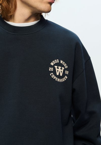Sweatshirt bleu marine avec un logo circulaire crème portant "WOOD WOOD COPENHAGUE" et l'année 2002. Col rond côtelé et texture douce.