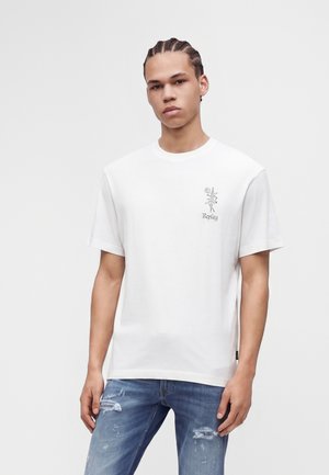Jeune homme aux cheveux tressés portant un t-shirt blanc avec un petit motif floral et texte, associé à un jean bleu déchiré sur un fond uni.