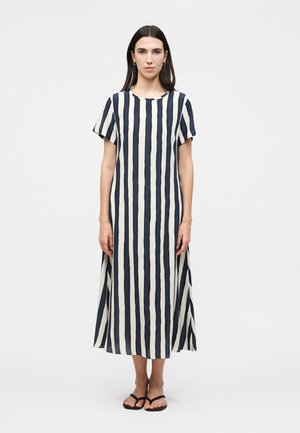 Γυναίκα με ίσια σκούρα μαλλιά που φοράει ένα κοντομάνικο φόρεμα μέχρι τον αστράγαλο με κάθετες μαύρες και άσπρες ρίγες και μαύρα σανδάλια, στέκεται με το πρόσωπο προς τα εμπρός.