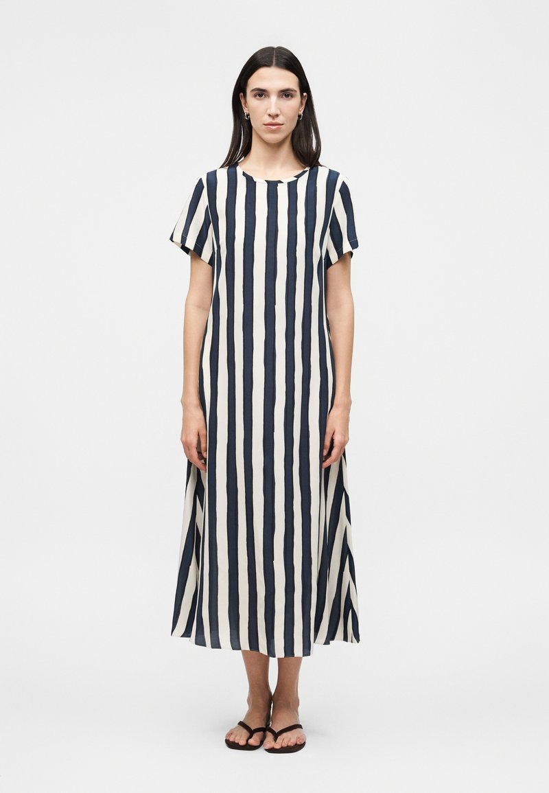 Γυναίκα με ίσια σκούρα μαλλιά που φοράει ένα κοντομάνικο φόρεμα μέχρι τον αστράγαλο με κάθετες μαύρες και άσπρες ρίγες και μαύρα σανδάλια, στέκεται με το πρόσωπο προς τα εμπρός.