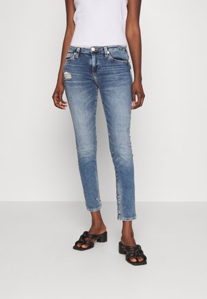 Kvinde iført lyseblå slidte skinny jeans, hvid ærmeløs top og sorte hælede sandaler med sammenknyttede stropper, stående mod hvid baggrund.