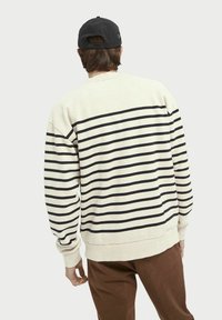 Sudadera a rayas beige con franjas horizontales negras. Puños y dobladillo acanalados, corte relajado. Llevada con pantalones marrones y una gorra negra.