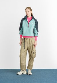 Πολυεστερικό πουλόβερ σε τιρκουάζ με μπλε μανίκια, μισό φερμουάρ και ροζ εσωτερικό γιακά; συνδυασμένο με χαλαρές μπεζ cargo παντελόνες με πλαϊνές τσέπες.