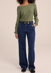 Haut à manches longues vert olive, jean ample bleu foncé, ceinture noire avec boucle dorée, baskets noires. Design simple avec une coupe décontractée.