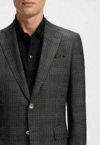 Ciemnozielona marynarka w kratę z wyraźnymi ramionami, zapinana na dwa guziki, z kieszonką na piersi trzymającą złożoną ciemnozieloną chusteczkę w kieszeni, w połączeniu z czarną koszulą.