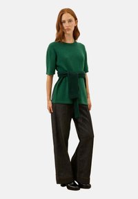 Zielony, krótki sweter z wiązaniem w talii, zestawiony z ciemnymi szerokimi dżinsami. Materiał wydaje się miękki, o teksturowanym wykończeniu.