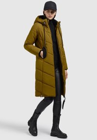 Abrigo acolchado de color verde oliva con capucha, cierre frontal con cremallera y bolsillos laterales, combinado con leggins texturizados negros y botas negras hasta la rodilla.