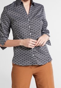 Chemise noire et blanche à boutons avec un motif floral géométrique, à manches longues et à coupe ajustée. Associée à un pantalon marron.