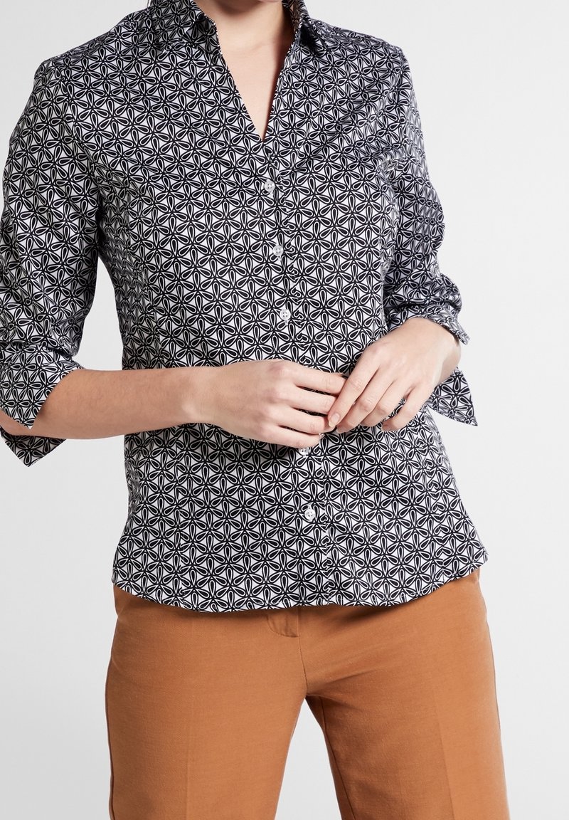 Chemise noire et blanche à boutons avec un motif floral géométrique, à manches longues et à coupe ajustée. Associée à un pantalon marron.