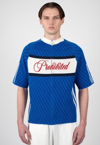 Joven con una camisa azul de manga corta con cremallera y la palabra "Prohibido" en escritura roja sobre un panel blanco en el pecho.