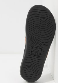 Zwarte rubberen schoenenzool met een golvend loopvlakpatroon. Bevat een rechthoekig logo met de opschrift "REEF" en een bergenafbeelding.