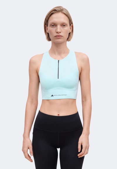 Frau trägt hellblaues ärmelloses Crop-Top mit Frontreißverschluss und schwarze hoch taillierte Leggings, steht vor einfarbigem Hintergrund.