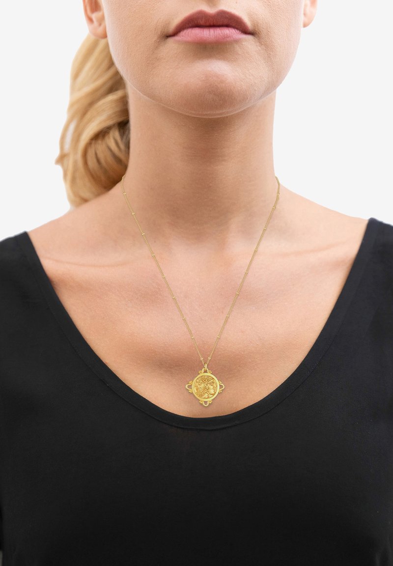 Collana d'oro con un pendente circolare caratterizzato da dettagli intricati, indossata su una maglietta nera con scollatura a V. La catena è delicata con una texture minima.