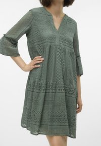 Robe en dentelle verte avec un décolleté en V, des manches trois-quarts et un design à volants. Présente des motifs élaborés et un matériau léger et texturé.