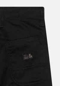 Juodi džinsai turi užpakalinį kišenę su siuvinėtu kontūru ir audinio etikete, ant kurios užrašyta "JACK & JONES", demonstruojantys subtilų tekstūrą ir detales.