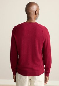 Roter Wollpullover mit Rundhalsausschnitt und langen Ärmeln; glatte Textur und gerippte Akzente an den Bündchen und am Saum. Minimalistisches Design.
