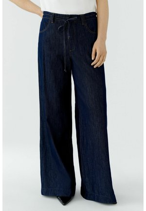 Frau trägt dunkelblaue Jeans mit weitem Bein und Kordelzug an der Taille sowie schwarze spitze Schuhe vor einfarbigem Hintergrund.