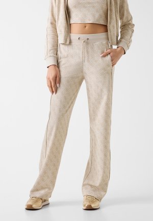 Femme portant un pantalon de survêtement beige à motifs à jambes larges avec taille à cordon, une veste courte zippée assortie et des baskets beiges.