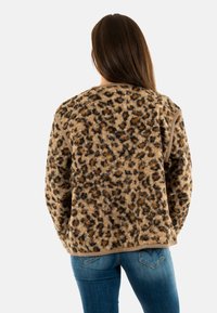 Donna che indossa una giacca di pile leopardata a maniche lunghe e jeans blu, voltata di spalle rispetto alla fotocamera su uno sfondo bianco.