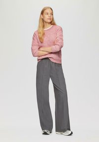Hellrosa strukturiertes Sweatshirt mit langen Ärmeln, kombiniert mit lockeren grauen Hosen. Das Model steht mit verschränkten Armen und trägt metallische Sneakers.