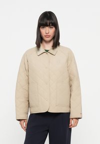Beige quiltet jakke med kragehals, store frontlommer og logoaccent på venstre brystområde. Myk stofftekstur.