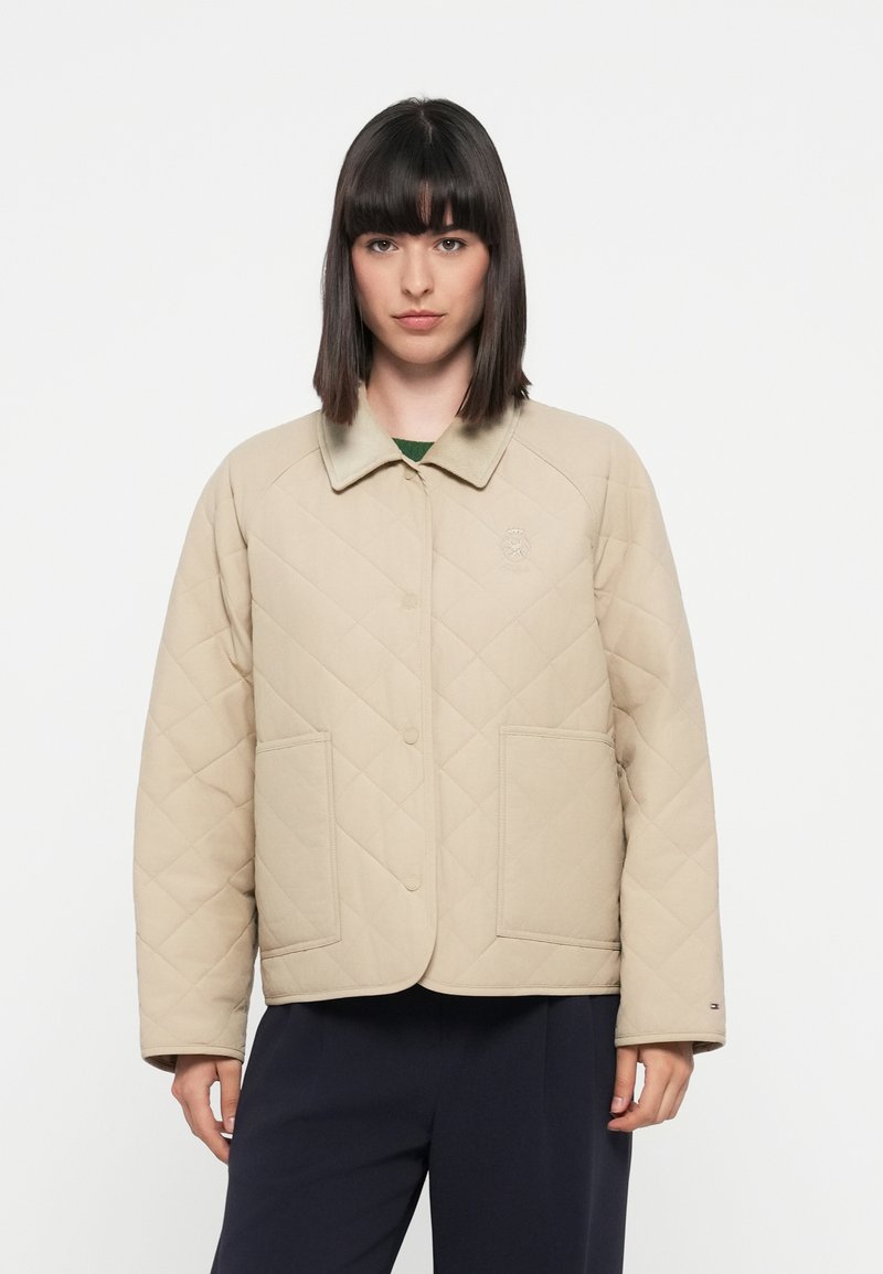 Beige quiltet jakke med kragehals, store frontlommer og logoaccent på venstre brystområde. Myk stofftekstur.
