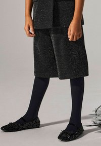 Črne bleščeče kratke hlače so v paru z mornarsko modrimi nogavicami in okrašenimi črnimi balerinkami. Material ima sijočo, odbojno teksturo.