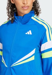 Chaqueta deportiva azul con acentos en blanco y verde neón, cierre completo, cuello alzado y detalles de panel lateral transpirable.