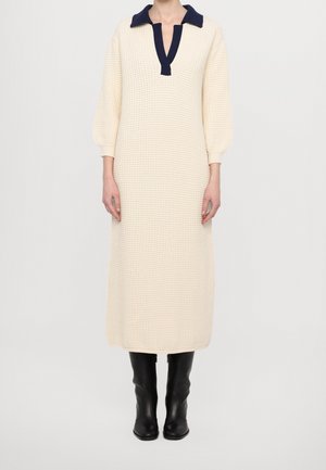 Femme portant une longue robe en maille crème avec un col bleu marine et des bottes noires montant jusqu'au genou, debout devant un fond clair uni.
