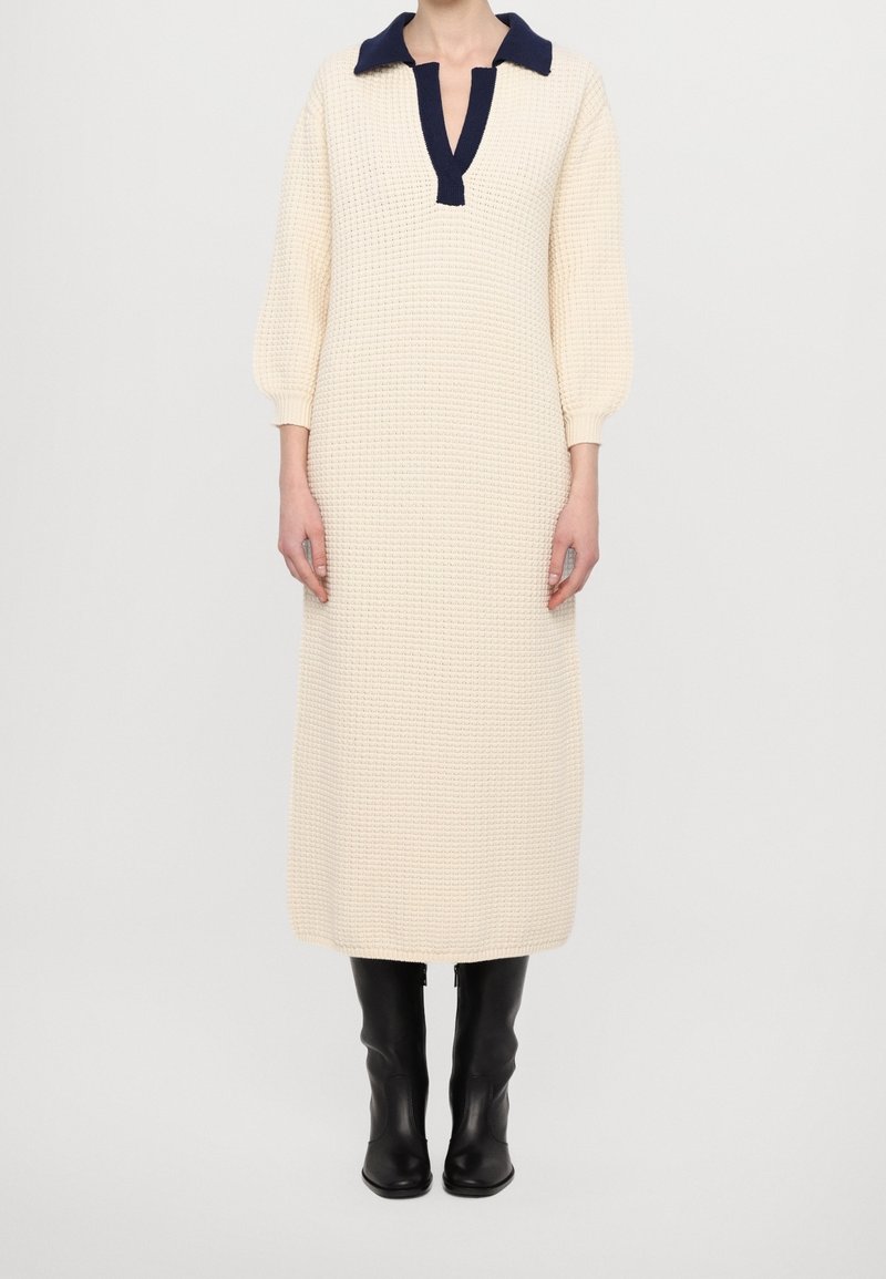 Femme portant une longue robe en maille crème avec un col bleu marine et des bottes noires montant jusqu'au genou, debout devant un fond clair uni.