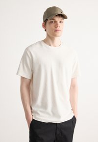 Beige t-shirt med kort ärm och rund halsringning, tillverkad av mjukt tyg. Kombinerad med en ljus olivgrön baseballkeps. Modellen poserar avslappnat.