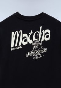 Sort skjorte med hvid tekst "Matcha Since 1987" og en tegneseriefigur af en matcha-drik, der holder et skilt med teksten "AMBASSADOR."