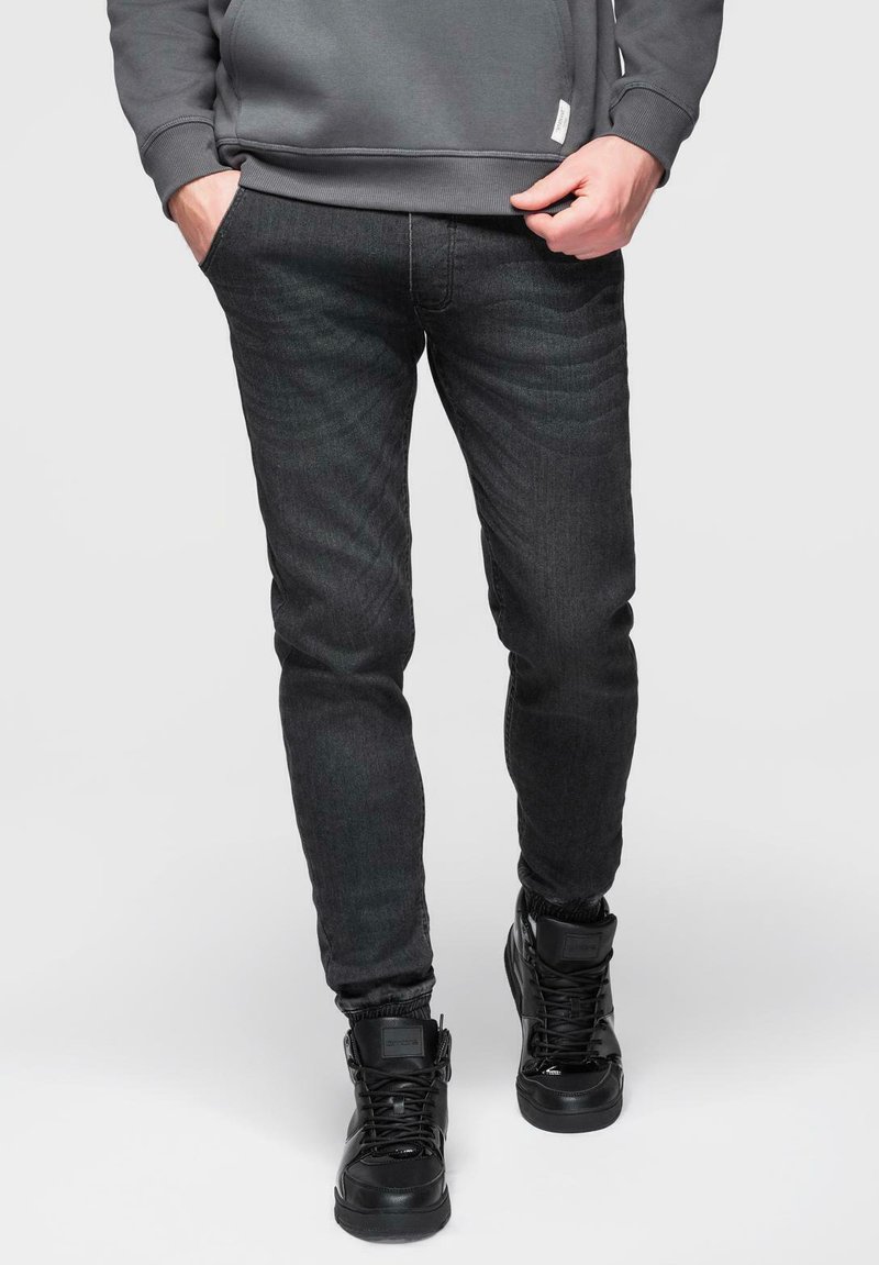 Jean noir slim avec une texture subtile, porté avec des baskets montantes noires et un sweat-shirt gris. Détails de couture minimes visibles.