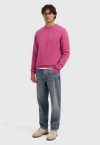 Rosa Sweatshirt mit Rundhalsausschnitt und gerippten Bündchen, kombiniert mit ausgewaschenen blauen Jeans. Das Outfit wird mit beigen Sneakers abgerundet.