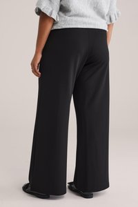 Mustat leveälahkeiset housut, jotka on valmistettu sileästä, joustavasta kankaasta. Niissä on siistit viimeistelyt ilman näkyviä taskuja. Yhdistetty mustiin tasapohjaisiin kenkiin.