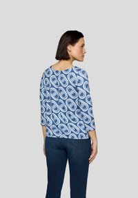 Bluse mit blauem Muster und dreiviertel Ärmeln, die kreisförmige Designs in Weiß und dunklerem Blau zeigt. Kombiniert mit dunkblauen Jeans.