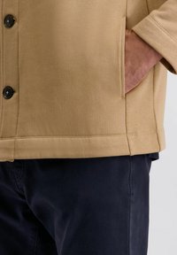 Tan katoenen jas met zwarte knopen, voorzien van een zak aan de zijkant en een relaxte pasvorm. Gecombineerd met donker marineblauwe broek.