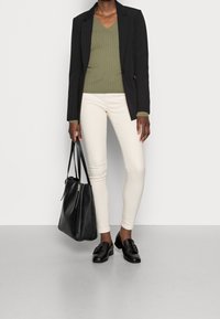 Svart blazer över en olivgrön ribbad tröja, krämfärgade skinny jeans och svarta loafers. Svart handväska med guldaccenter.