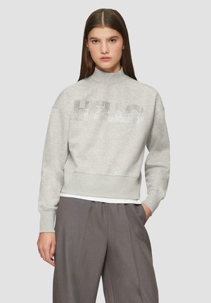 Light grey sweatshirt with high neckline, bold silver sequined "HELLO" text across the chest, and ribbed cuffs and hem. Worn over gray trousers.