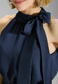 Gros plan sur une personne portant un haut sans manches bleu marine avec un détail en nœud au niveau du cou, mettant en valeur ses lèvres et une partie de son épaule.