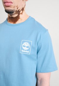 Világoskék pamut póló rövid ujjakkal, amelyen egy fehér Timberland logó található bal mellen, négyzet alakú keretben.