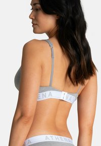 Soutien-gorge de sport gris avec des bretelles réglables, une bande blanche portant le mot "ATHENA" et une surface en tissu lisse. Fermeture au dos avec un crochet et un œillet.