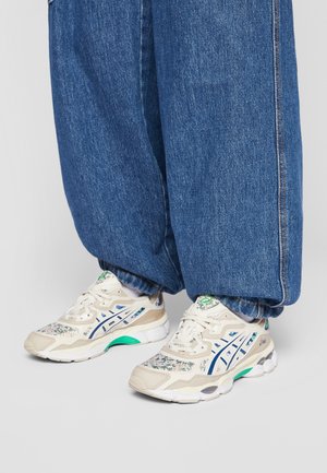 Person wearing loose blue denim jeans and white sneakers with blue and green floral patterns, standing on a light gray floor.