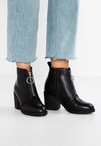 Bottes noires à la cheville en cuir lisse avec une fermeture éclair latérale et des détails ronds en argent, dotées d'un talon haut épais et d'une semelle texturée.