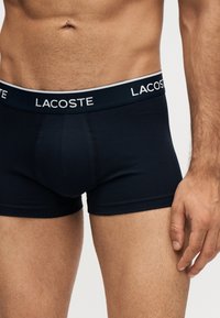 Αντρικός κορμός με μαύρα μποξεράκια Lacoste με εμφανές λογότυπο στην μέση και ασημένιο σκουλαρίκι στον αφαλό.