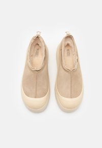 Beige Wildleder Slipper mit plüschiger Innenauskleidung, runder Zehenform und strukturiertem Gummi-Sohle. Enthält eine kleine Schlaufe auf der Rückseite für einfaches Anziehen.