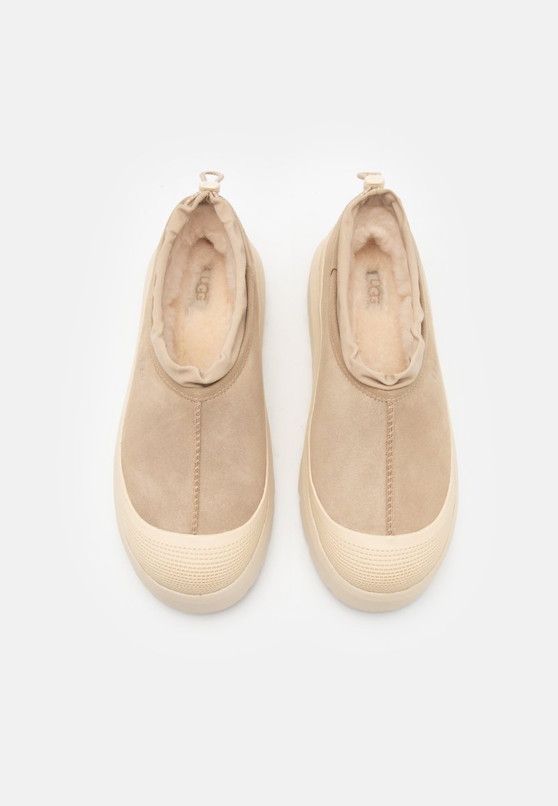 Chaussures en daim beige à enfiler avec une doublure en peluche, un bout rond et une semelle en caoutchouc texturé. Comprend une petite boucle à l'arrière pour un enfilage facile.