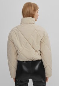 Beige quiltet jakke med høj krave, der har et diamantmønster og en cropped design. Båret over sorte slim-fit bukser.