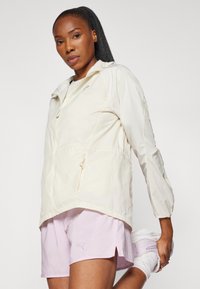 Helle beige Jacke mit Kordelzug in der Taille, durchgehendem Reißverschluss und zwei vorderen Taschen. Getragen mit rosa Shorts. Glatte Textur, lässiges Design.
