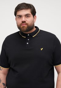 Čierne polo tričko vyrobené z textúrovaného materiálu, s žltým akcentom na golieri, trojbuttonovým zapínaním a malým žltým logom na hrudi.